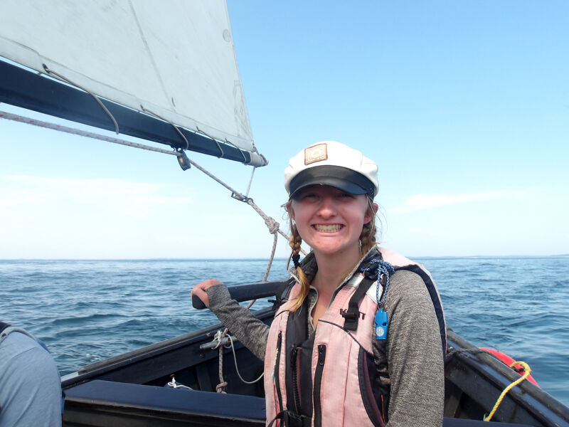 A woman is sailing on a sunny day. She is wearing a life jacket and a white cap. The sailboat is small and black, and the sail is white. The water is blue and there are some clouds in the sky. The woman is smiling and looks like she is enjoying herself.